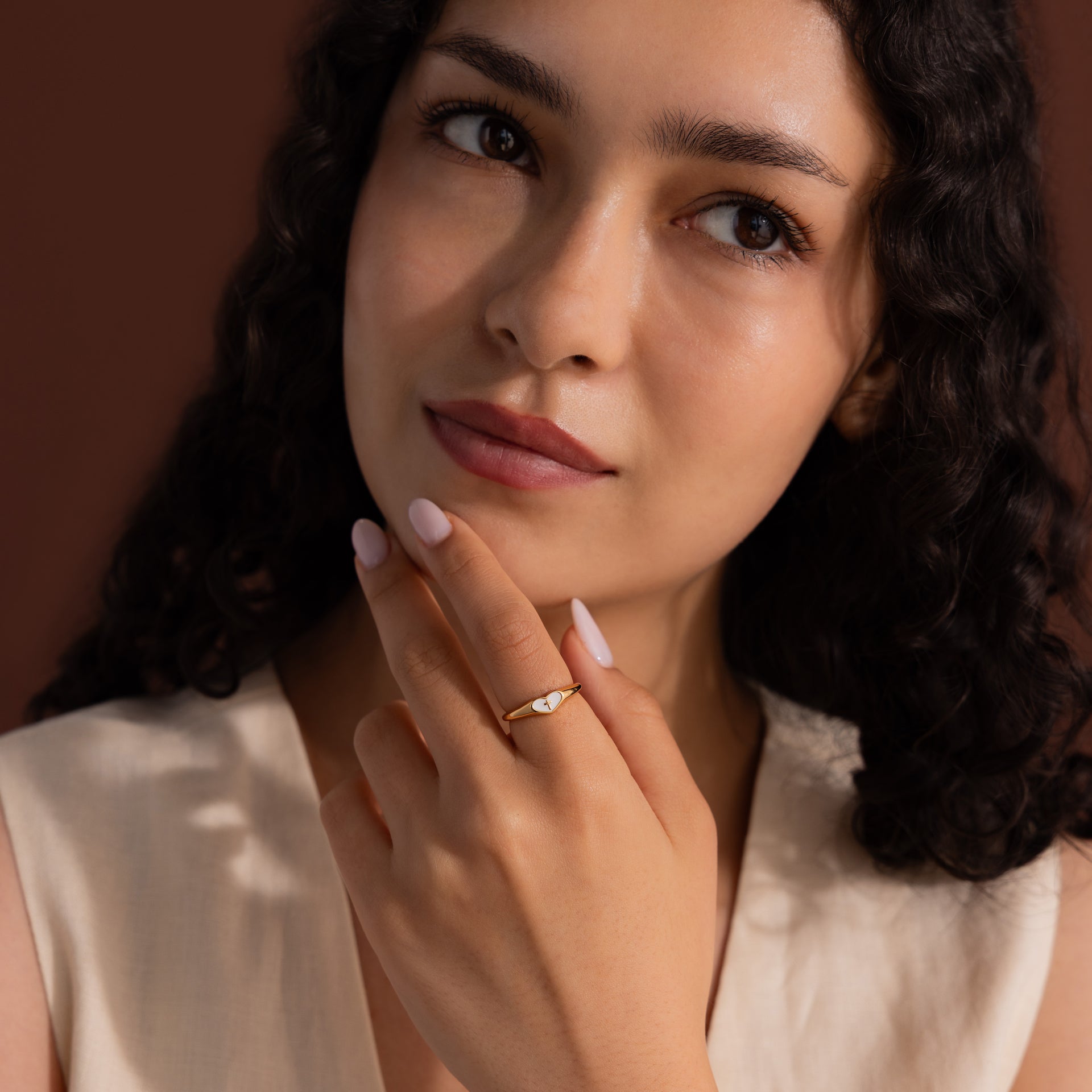 Model wearing a gold heart-shaped ring with a white enamel face and small gold cross.