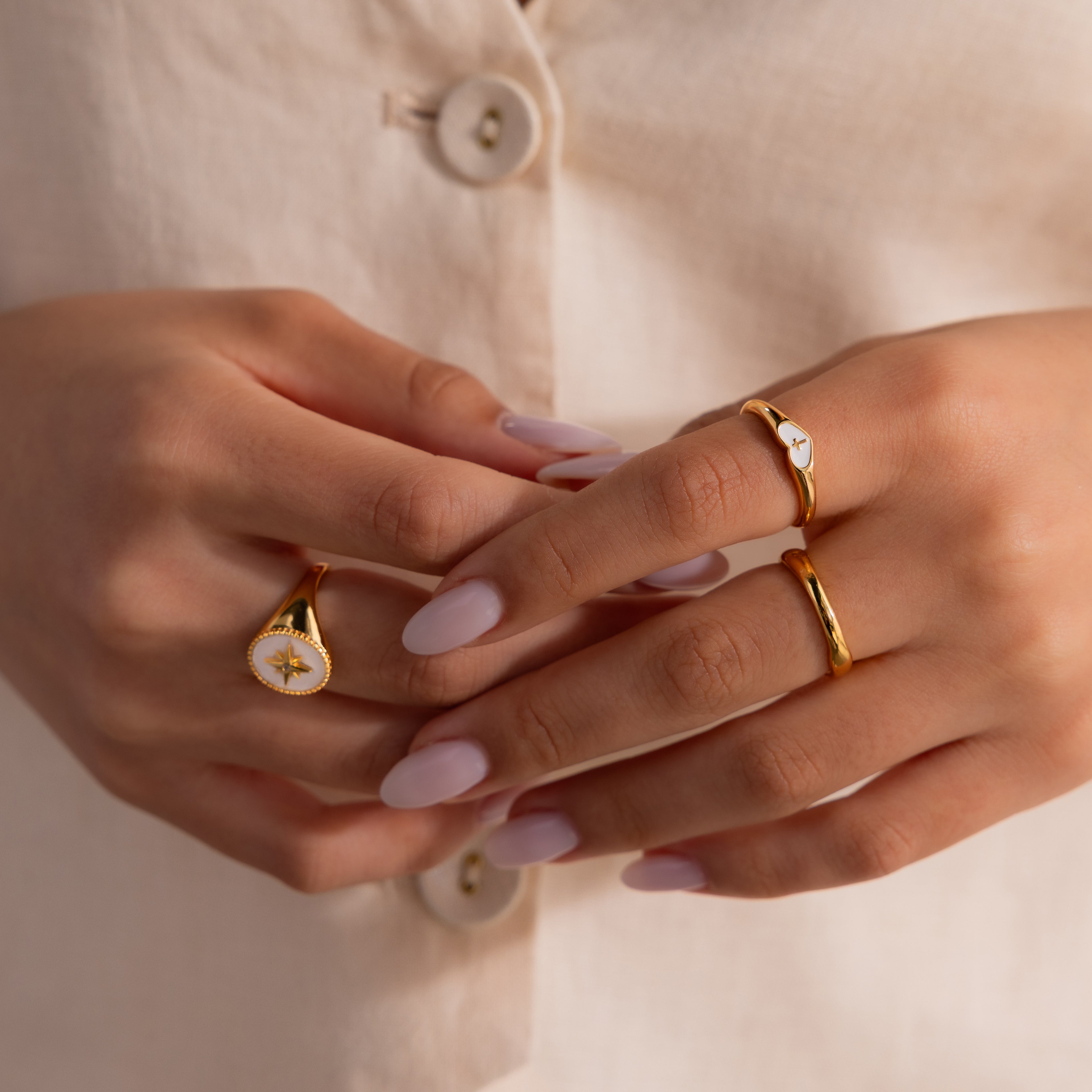Model wearing a gold heart-shaped ring with a white enamel face and small gold cross, stacked with multiple gold rings.
