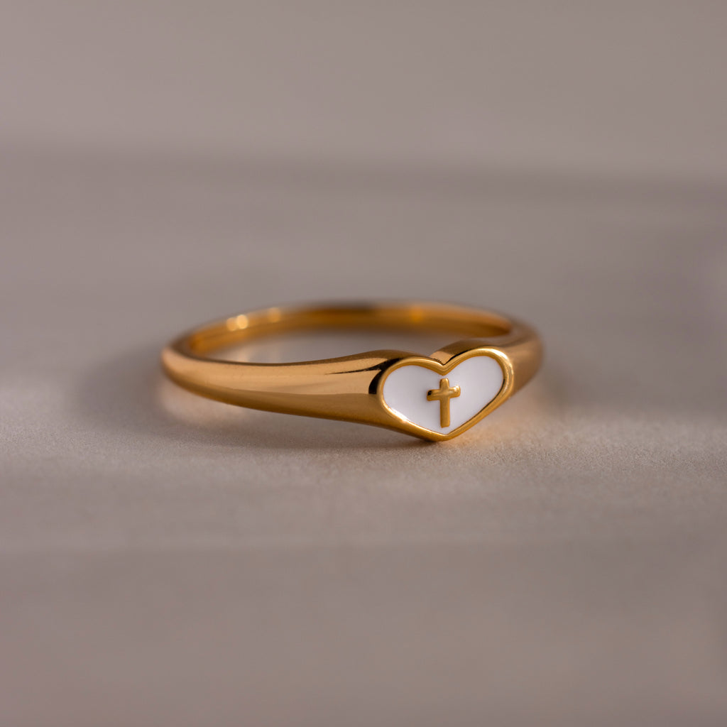 Close-up of a gold heart-shaped ring with white enamel face and small gold cross, displayed on a neutral surface.