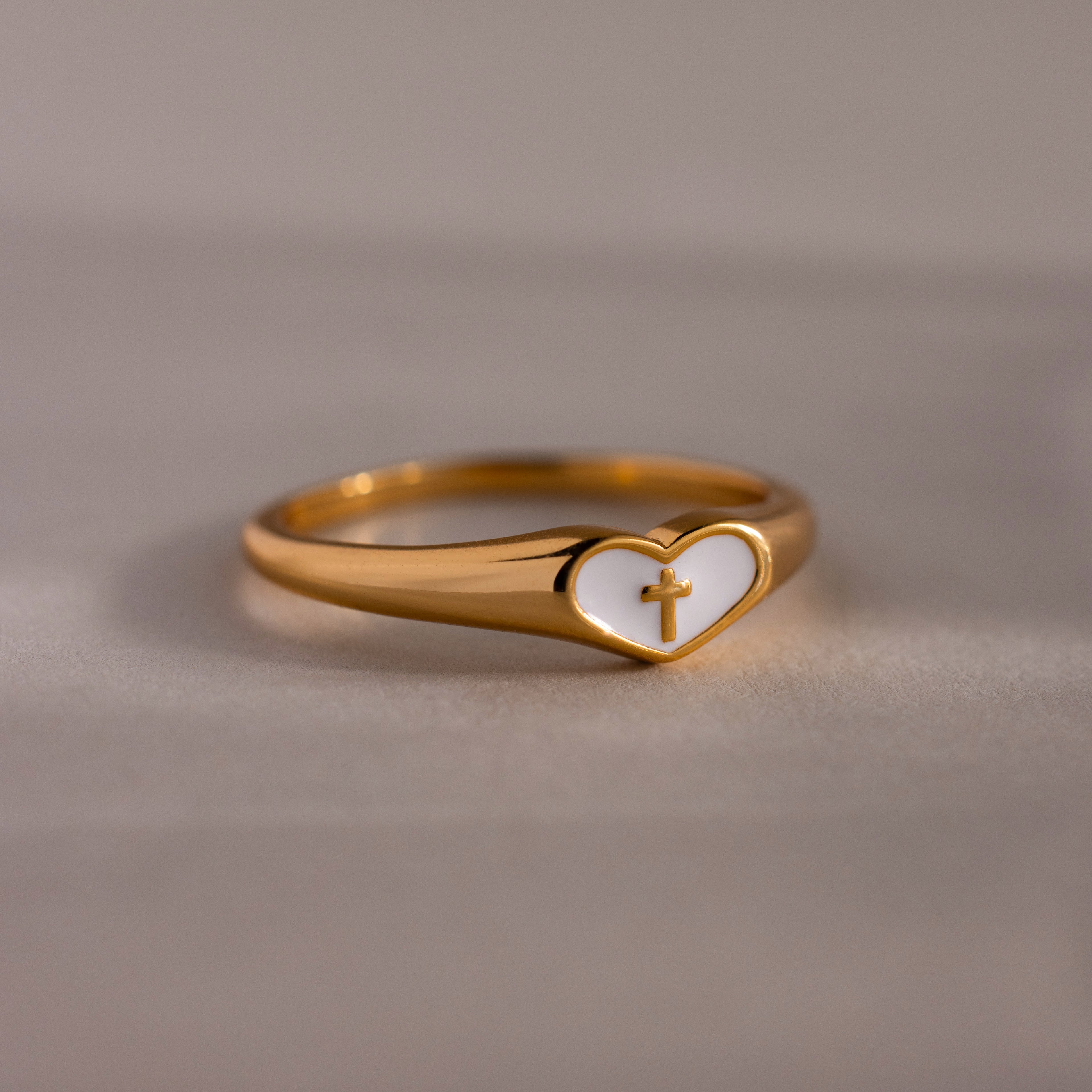 Close-up of a gold heart-shaped ring with white enamel face and small gold cross, displayed on a neutral surface.