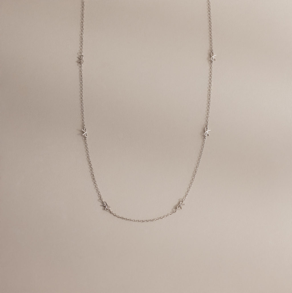 The Dainty Bow Necklace laid flat on a neutral surface, showing its full length, silver chain, and petite bow details.