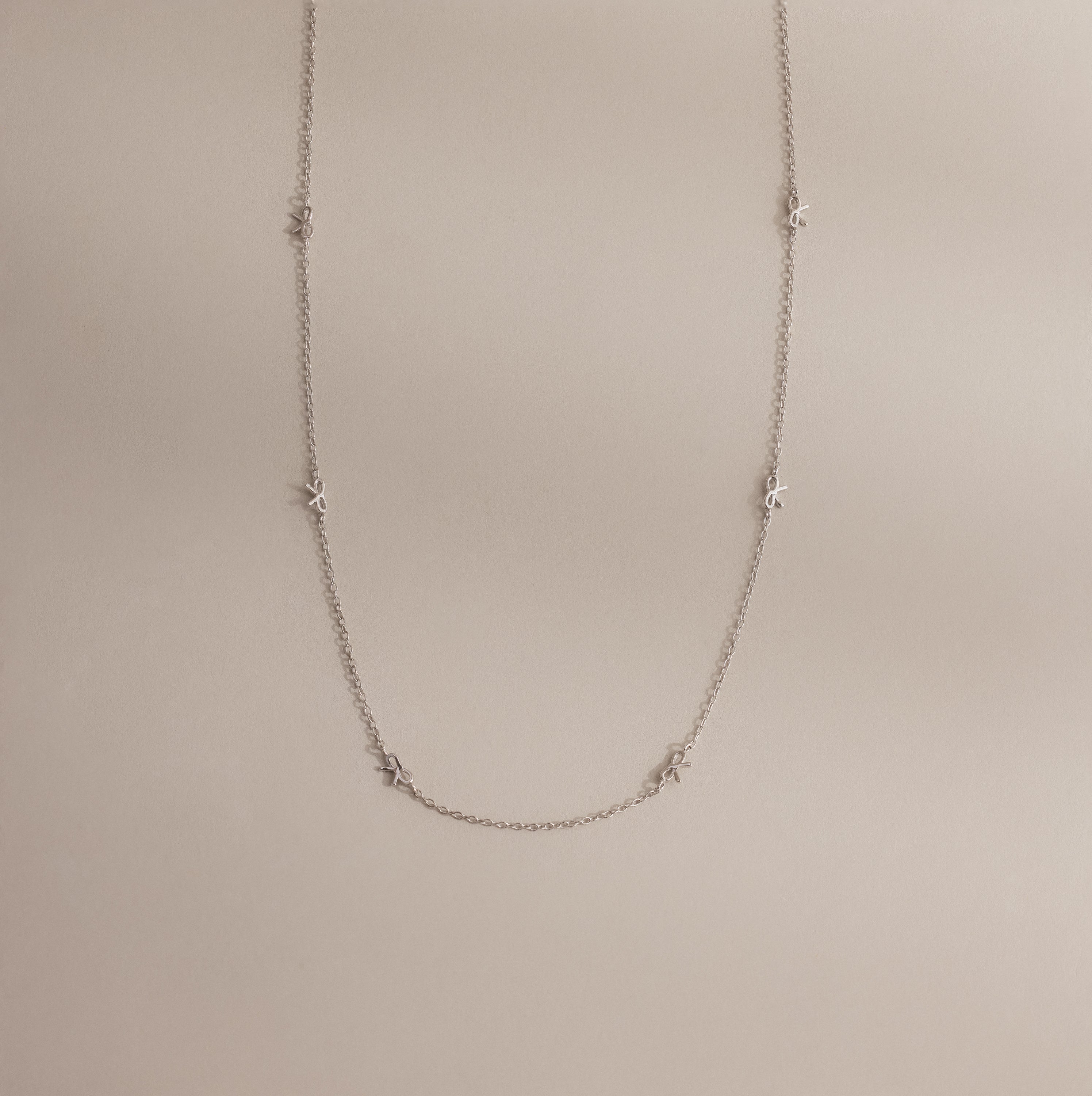 The Dainty Bow Necklace laid flat on a neutral surface, showing its full length, silver chain, and petite bow details.