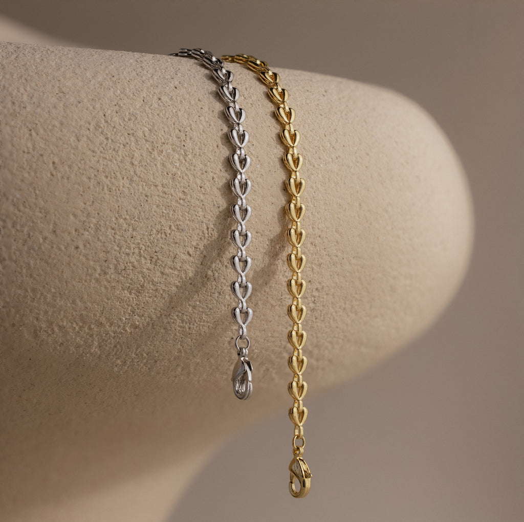 Side-by-side gold and silver Tiny Link Heart Bracelets draped over a neutral ceramic stand.