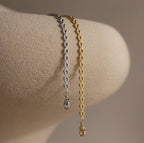 Side-by-side gold and silver Tiny Link Heart Bracelets draped over a neutral ceramic stand.