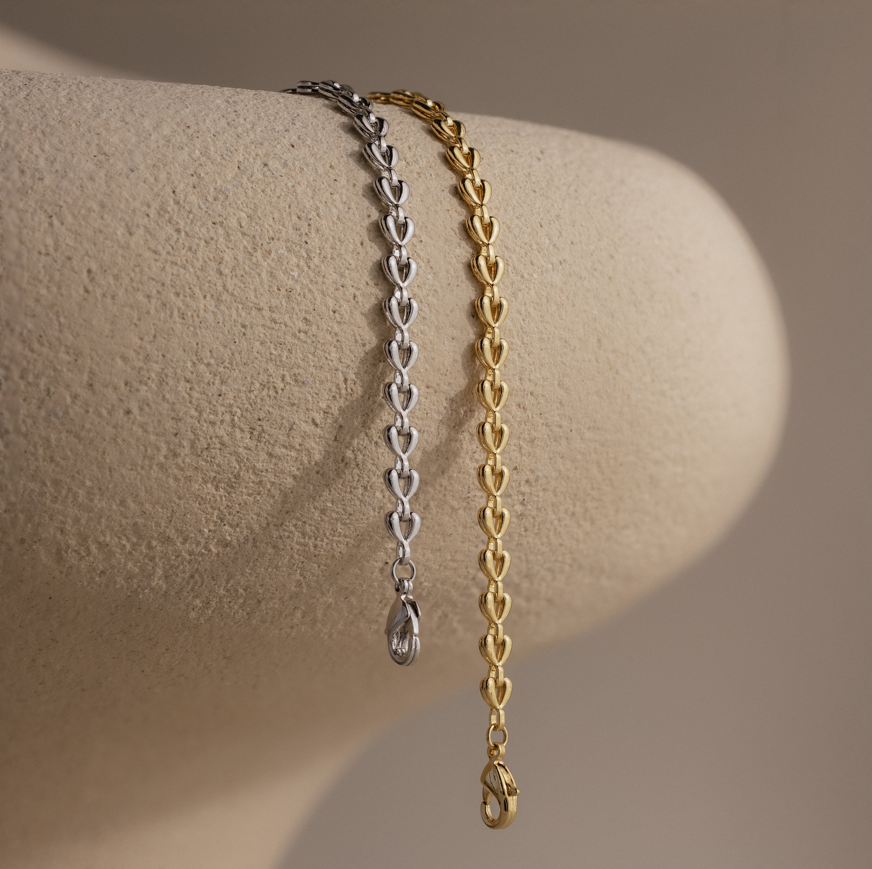 Side-by-side gold and silver Tiny Link Heart Bracelets draped over a neutral ceramic stand.