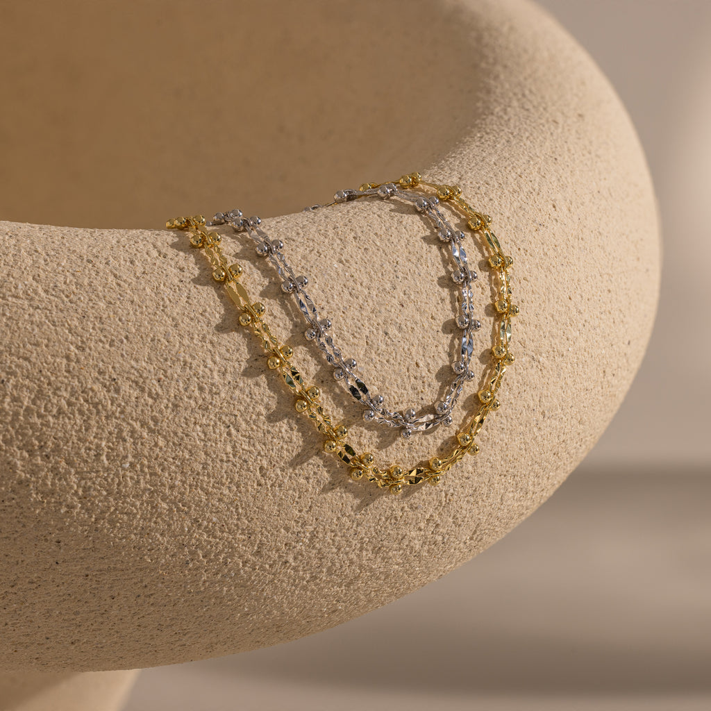 One gold and one silver Dainty Mirror Chain Bracelet draped over a textured ceramic surface.
