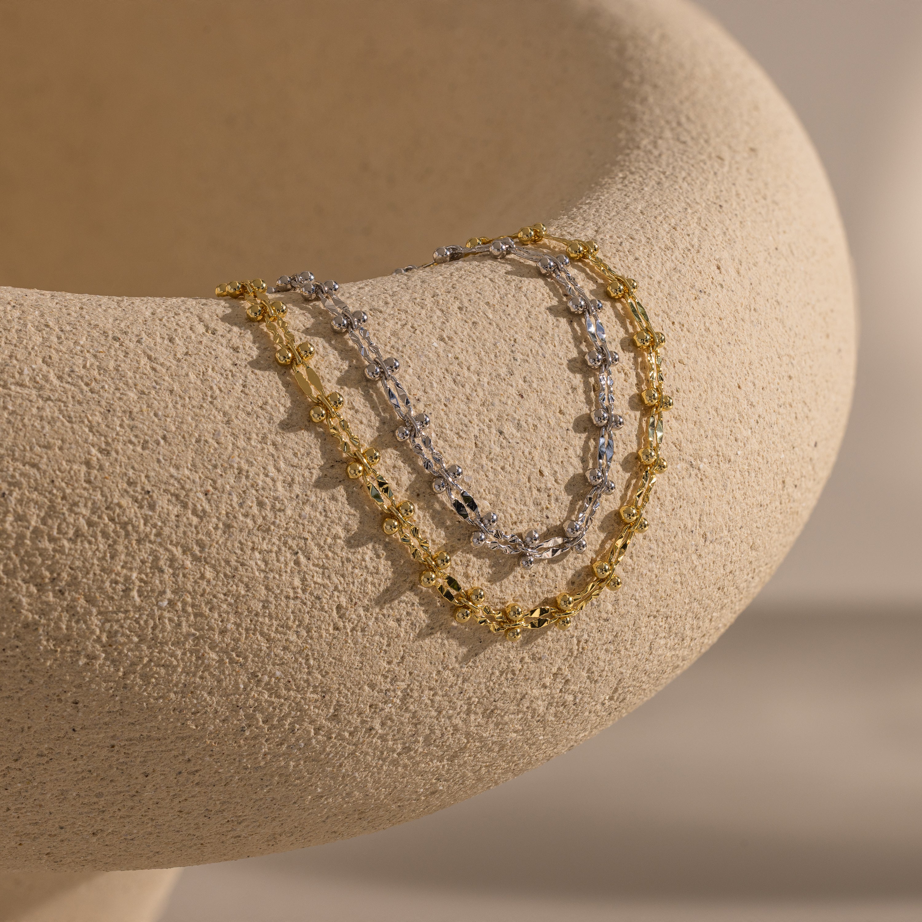 One gold and one silver Dainty Mirror Chain Bracelet draped over a textured ceramic surface.
