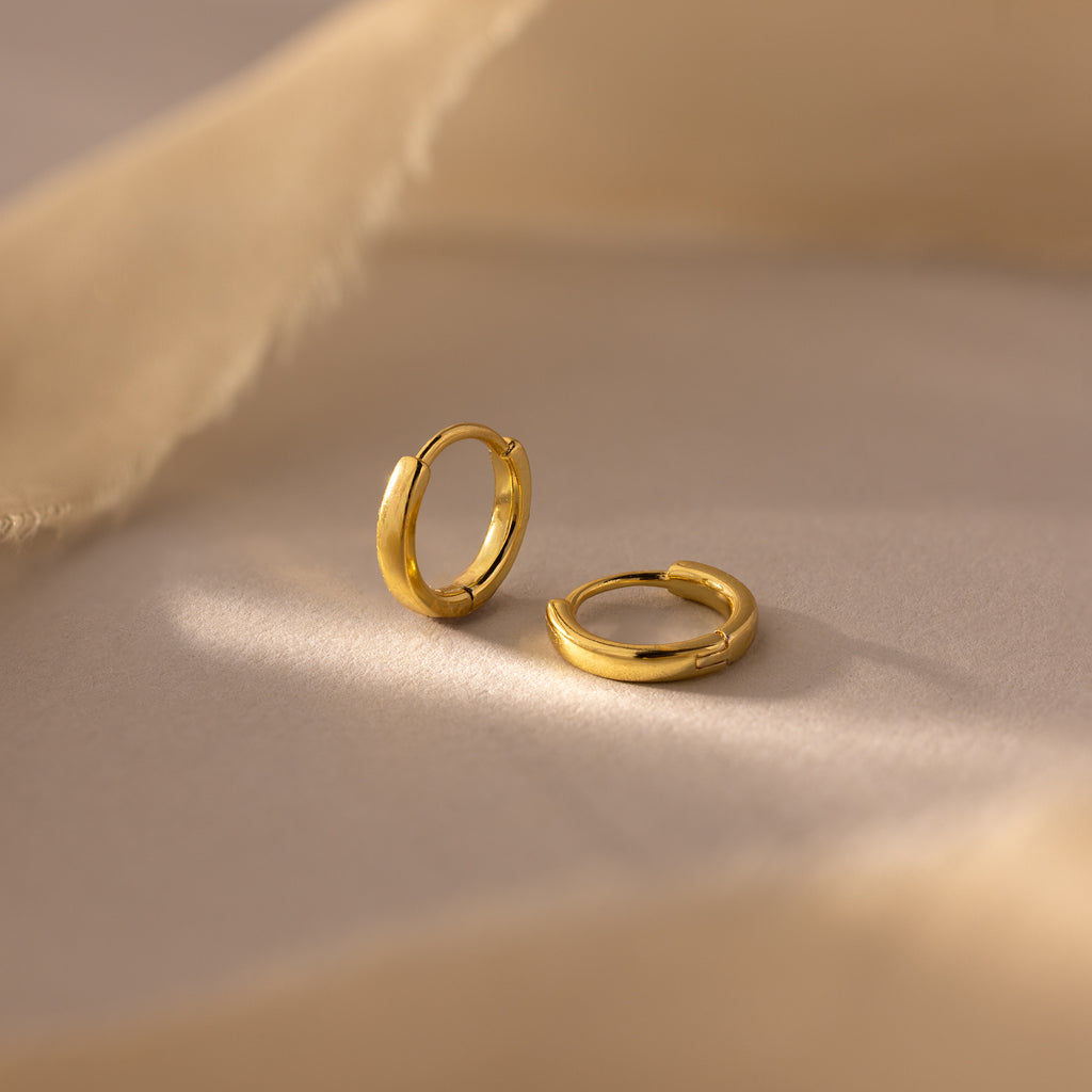 A pair of 7mm gold mini classic huggie hoop earrings displayed on a soft beige surface.