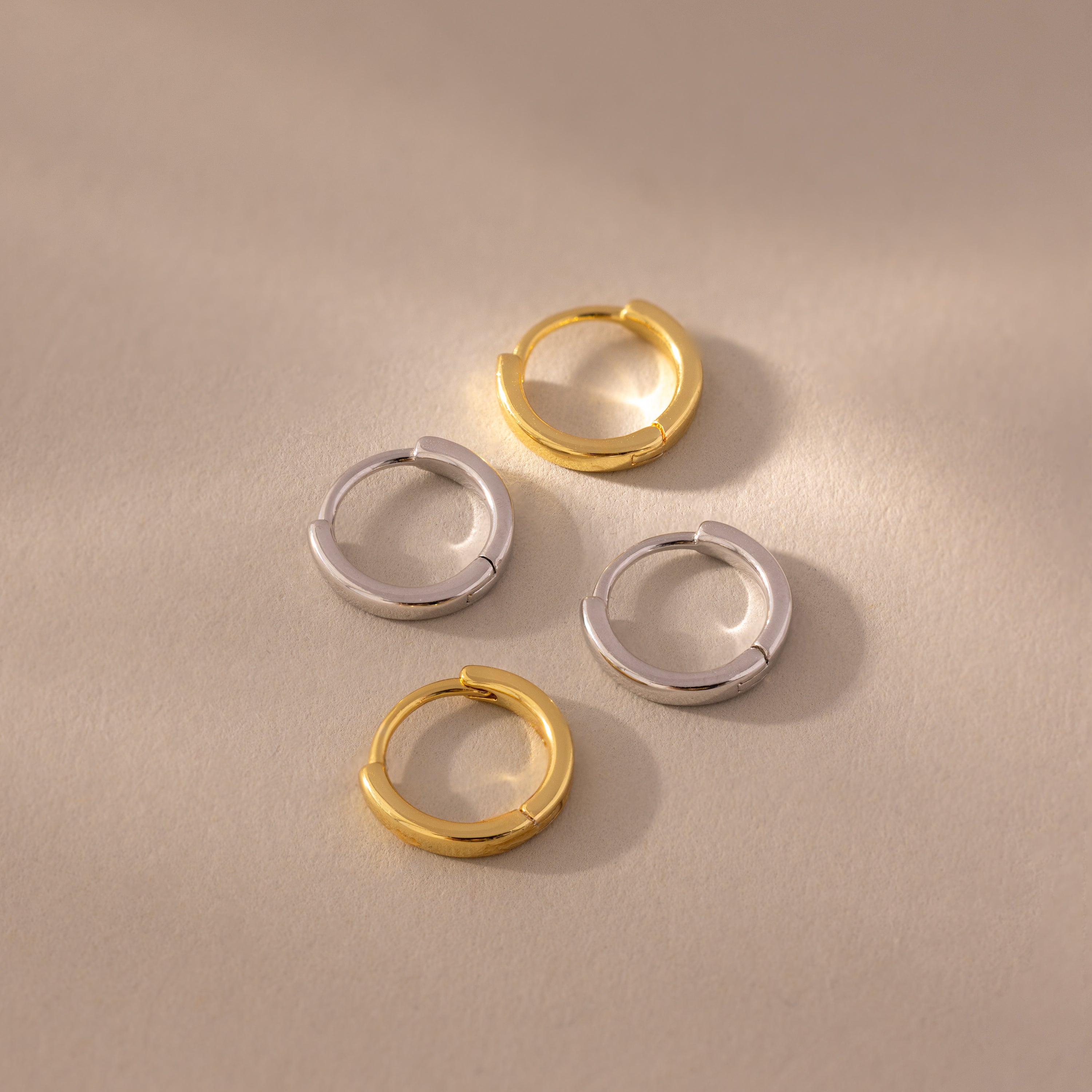 Flat lay of four 7mm Mini Classic Huggie Hoop Earrings in gold and silver finishes, styled on a neutral-toned surface with soft lighting.