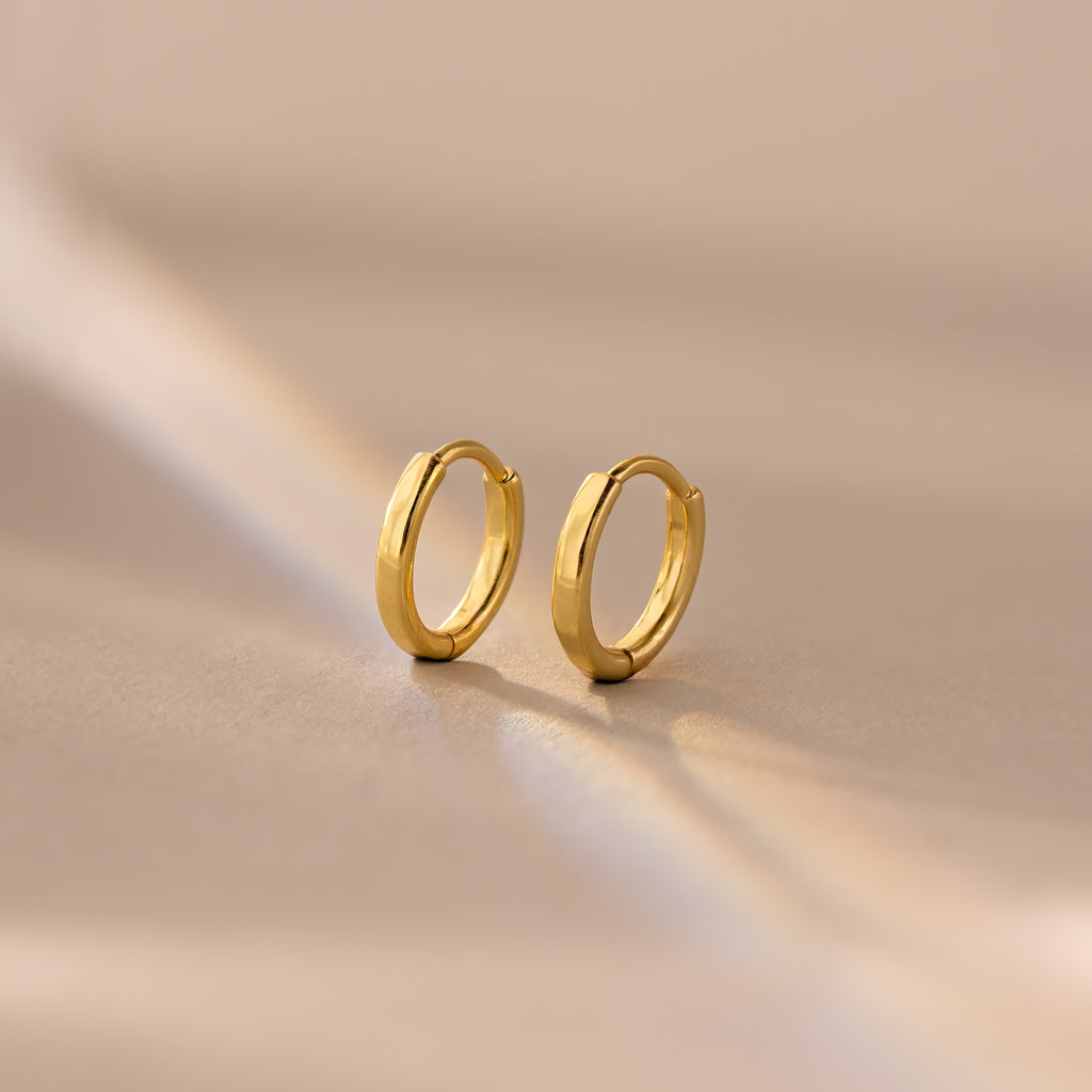 Pair of 8mm gold small dainty huggie hoop earrings resting on a soft beige surface.