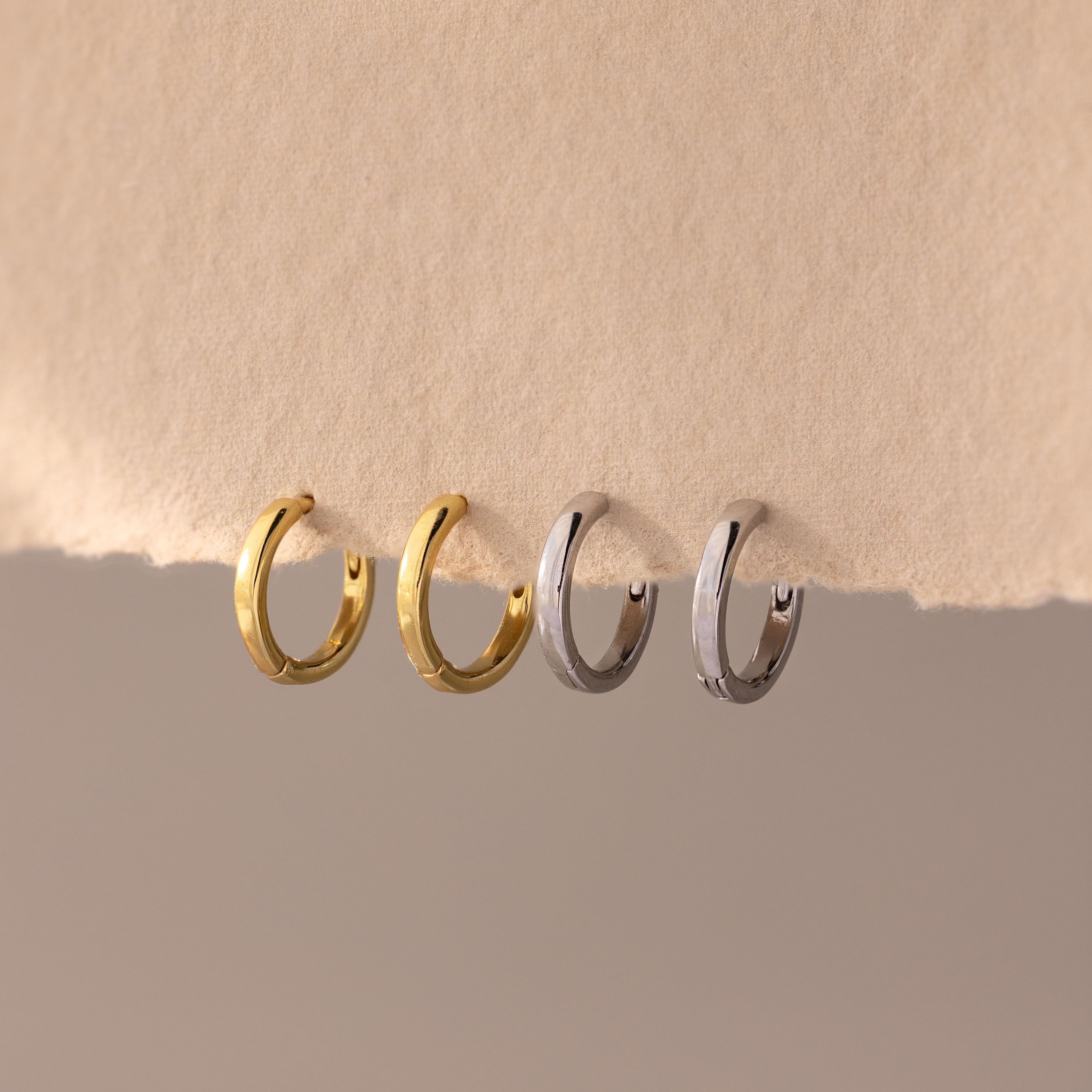 Set of four 8mm huggie hoop earrings in gold and silver finishes, displayed hanging on a textured neutral-toned paper edge.
