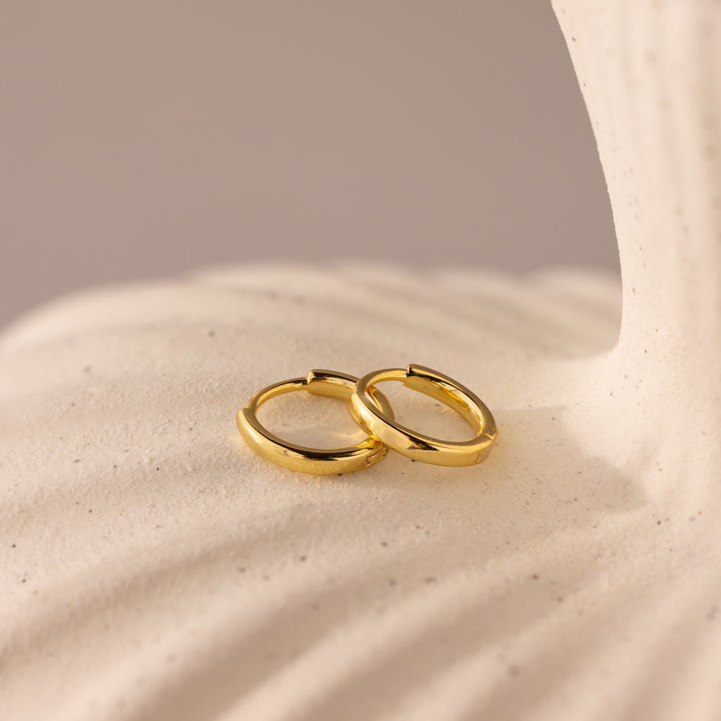 A pair of polished gold 8mm small classic huggie hoop earrings resting on a softly textured cream surface.