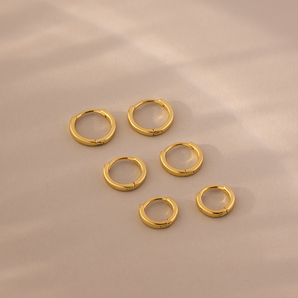 Flat lay of six gold huggie hoop earrings in three different sizes on a soft beige background.