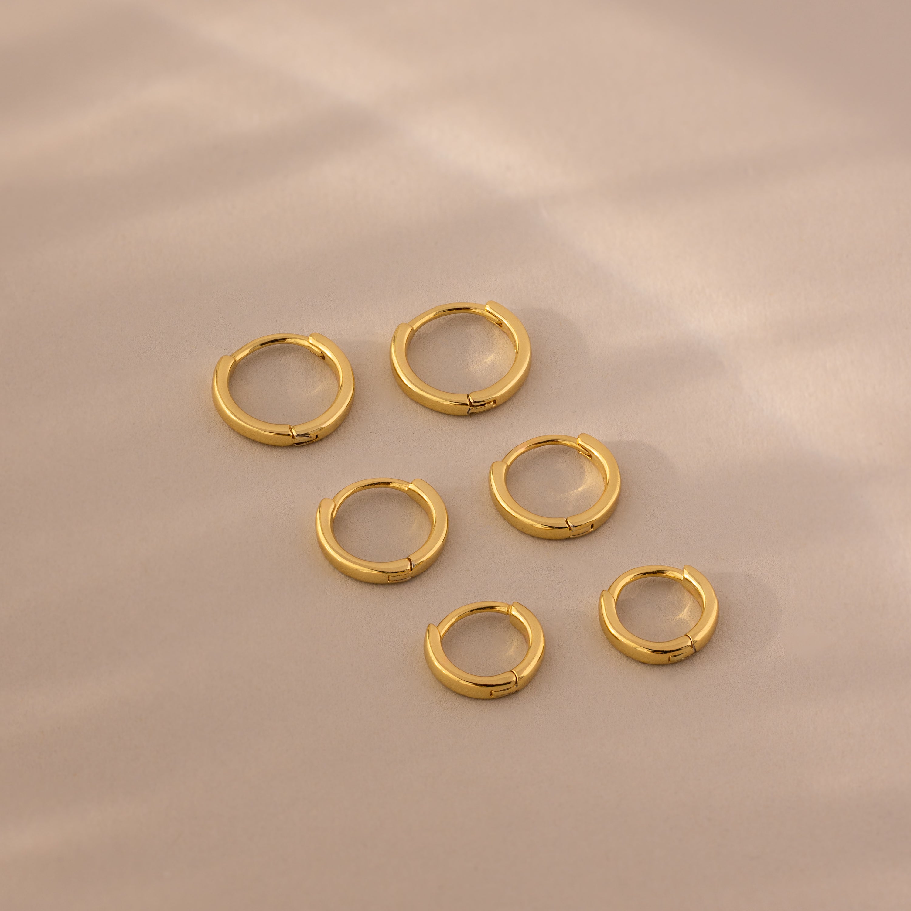 Flat lay of six gold huggie hoop earrings in three different sizes on a soft beige background.
