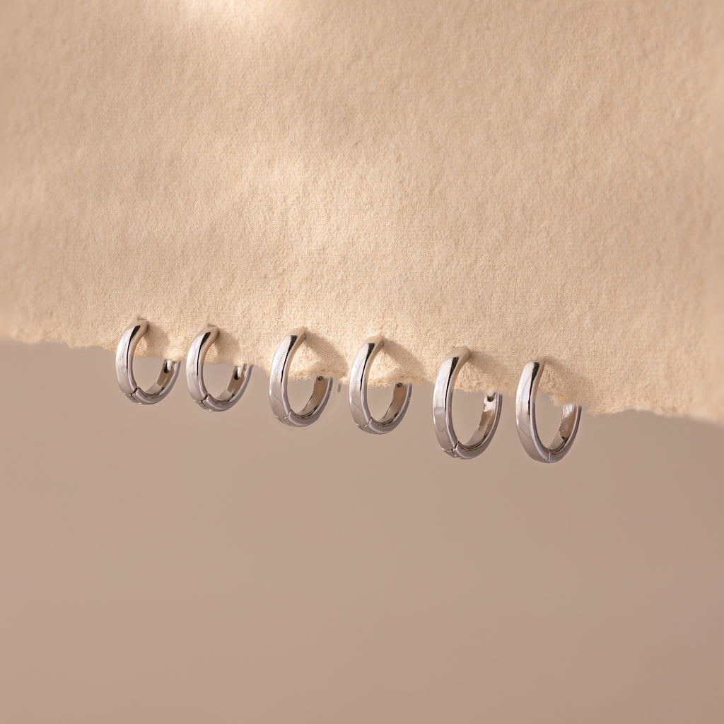 Row of six polished silver huggie hoop earrings in various sizes, neatly displayed on a soft beige textured fabric.