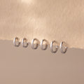 Row of six polished silver huggie hoop earrings in various sizes, neatly displayed on a soft beige textured fabric.