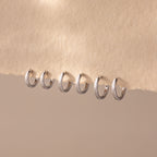 Row of six polished silver huggie hoop earrings in various sizes, neatly displayed on a soft beige textured fabric.
