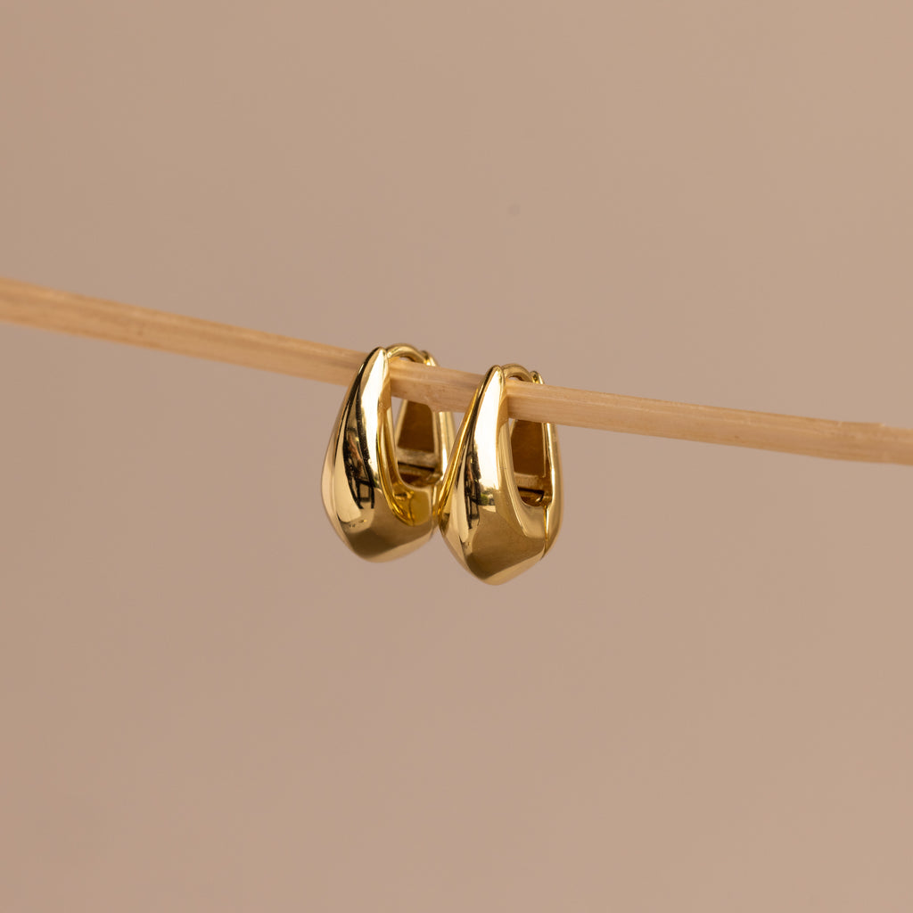 Pair of gold oval-shaped huggie earrings hanging delicately on a thin wooden rod against a neutral beige background.