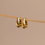 Pair of gold oval-shaped huggie earrings hanging delicately on a thin wooden rod against a neutral beige background.