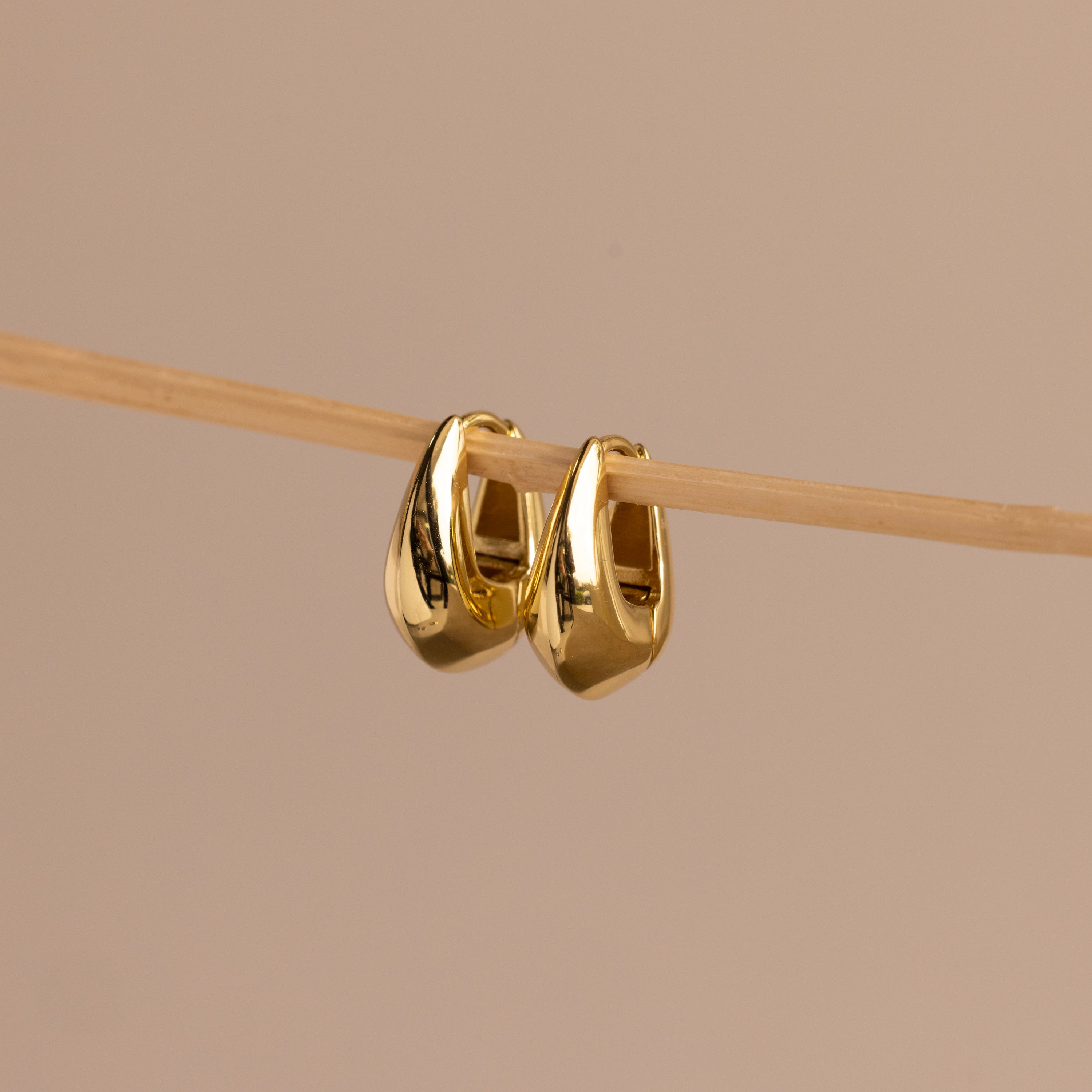 Pair of gold oval-shaped huggie earrings hanging delicately on a thin wooden rod against a neutral beige background.