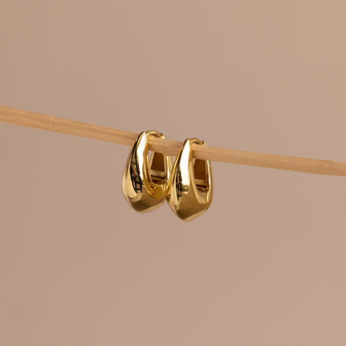 Pair of gold oval-shaped huggie earrings hanging delicately on a thin wooden rod against a neutral beige background.