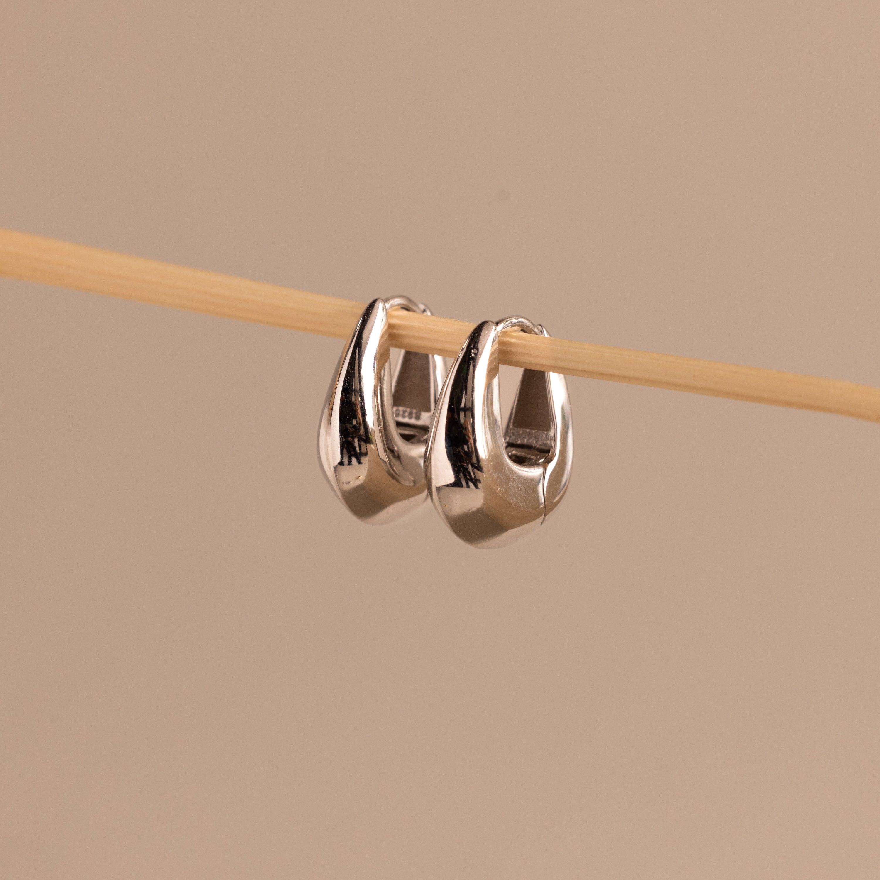 Close-up of a pair of polished silver oval-shaped huggie earrings hanging from a wooden rod.