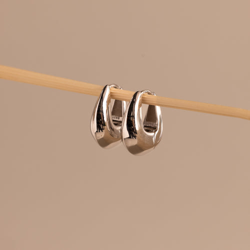 Close-up of a pair of polished silver oval-shaped huggie earrings hanging from a wooden rod.
