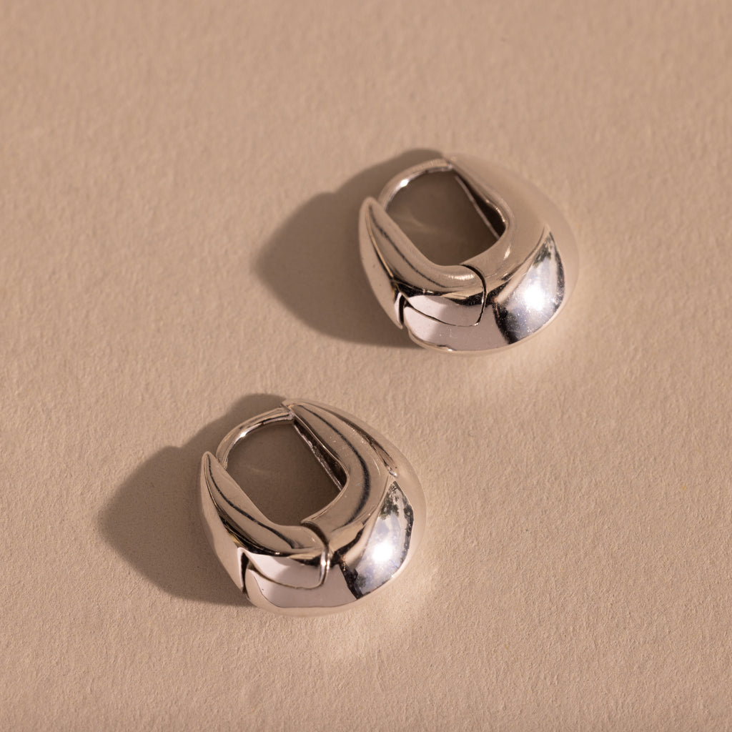 Pair of thick silver oval-shaped huggie earrings displayed flat on a neutral textured surface.