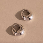 Pair of thick silver oval-shaped huggie earrings displayed flat on a neutral textured surface.
