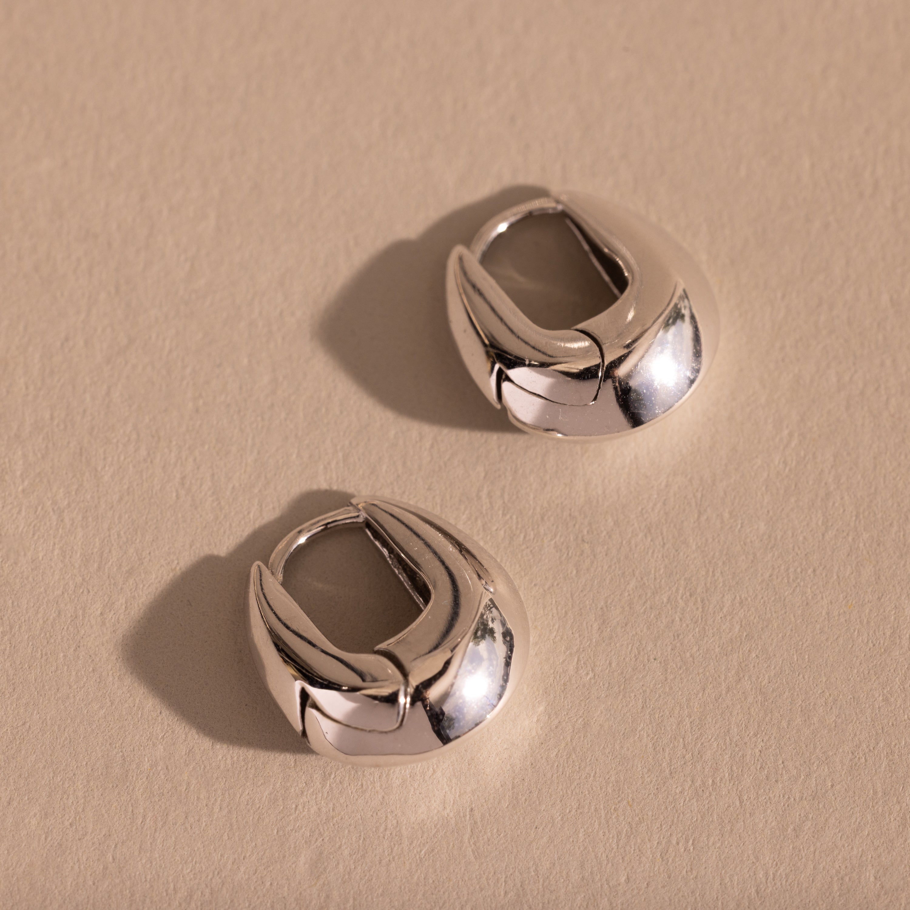 Pair of thick silver oval-shaped huggie earrings displayed flat on a neutral textured surface.