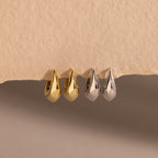 Four bold oval-shaped huggie hoop earrings in gold and silver finishes, displayed on a textured beige surface.