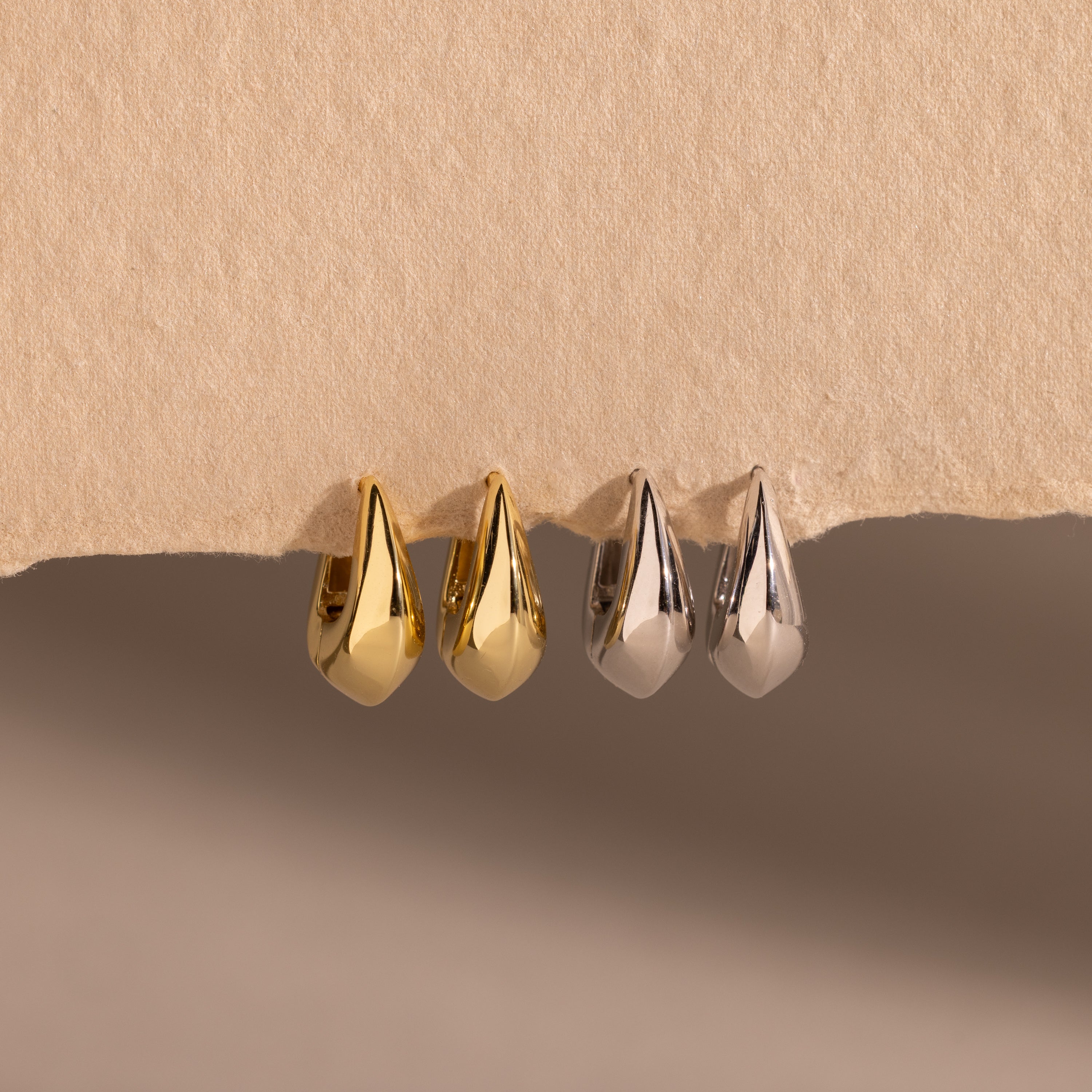 Four bold oval-shaped huggie hoop earrings in gold and silver finishes, displayed on a textured beige surface.