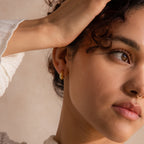 Model wearing a single gold oval-shaped huggie earring, with her hand gently resting on her head.