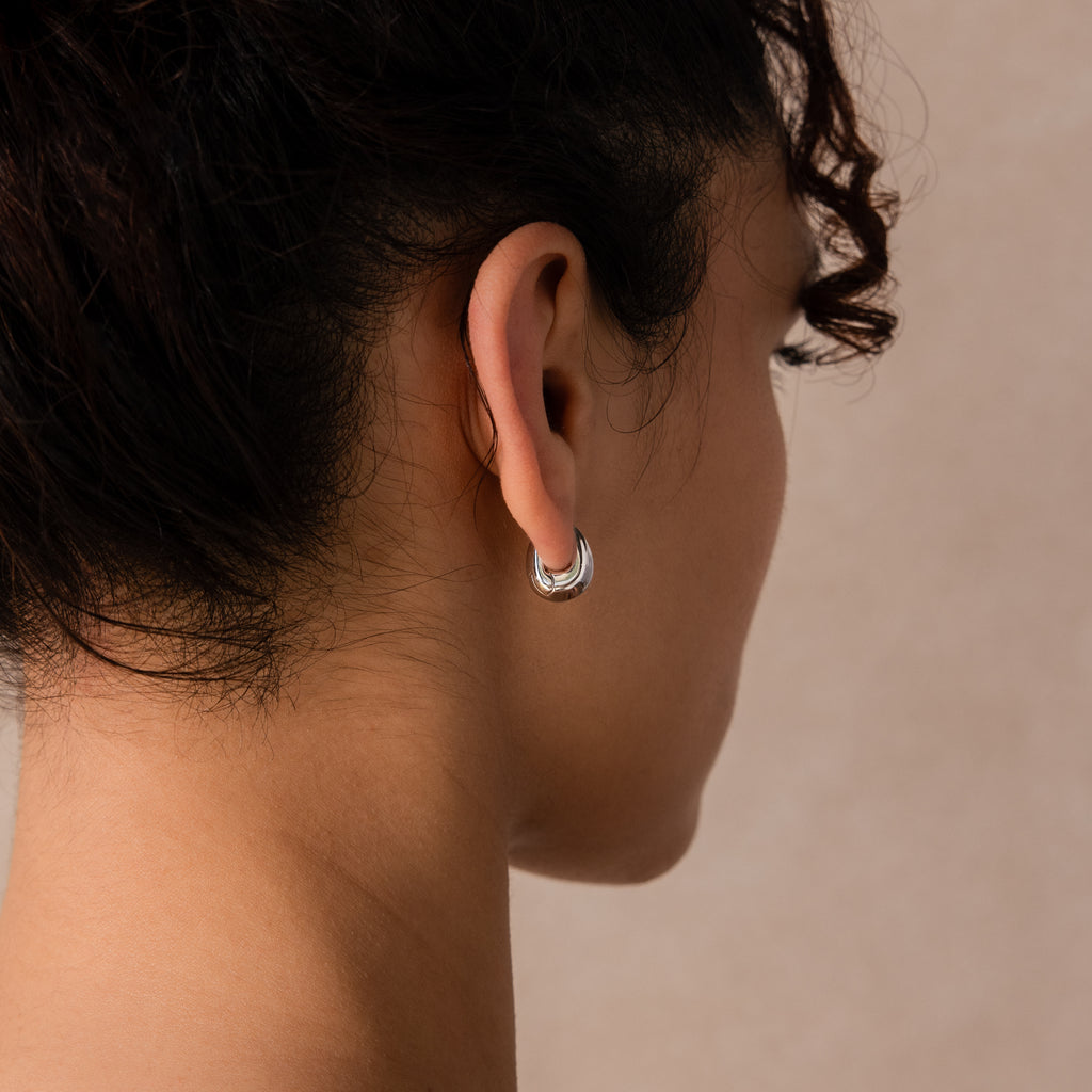 Close-up of model wearing a thick silver oval huggie earring against a neutral background.