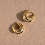 Pair of gold oval-shaped hoop earrings displayed flat on a neutral textured surface.