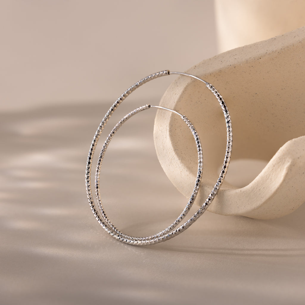 Close-up of large textured silver hoop earrings resting elegantly against a sculptural neutral-toned backdrop.