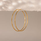 Close-up of large textured gold hoop earrings hanging against a soft, neutral-toned background.