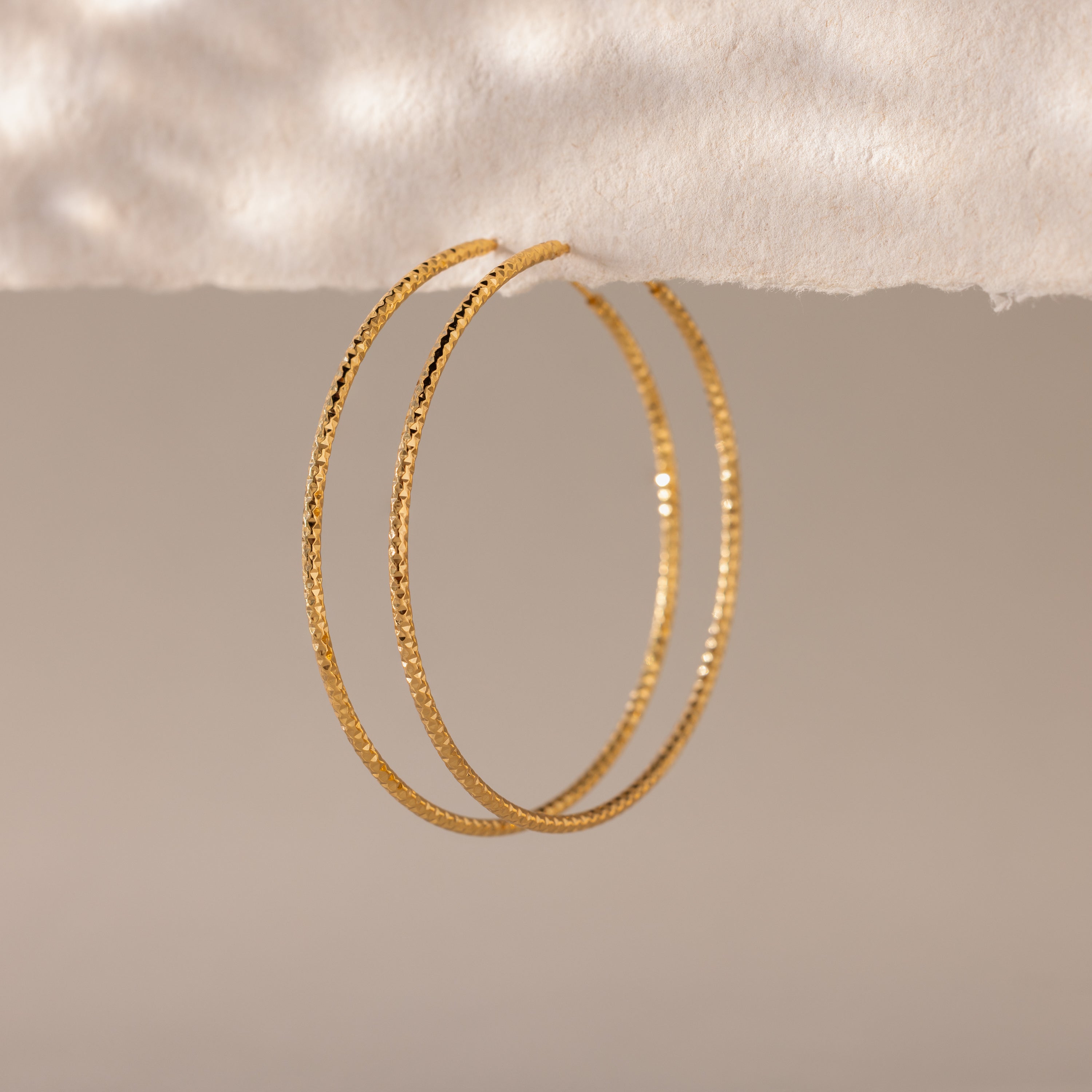 Close-up of large textured gold hoop earrings hanging against a soft, neutral-toned background.