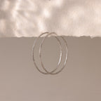 A pair of large, silver-toned hoop earrings with a faceted texture are suspended from the edge of a softly torn piece of off-white paper.