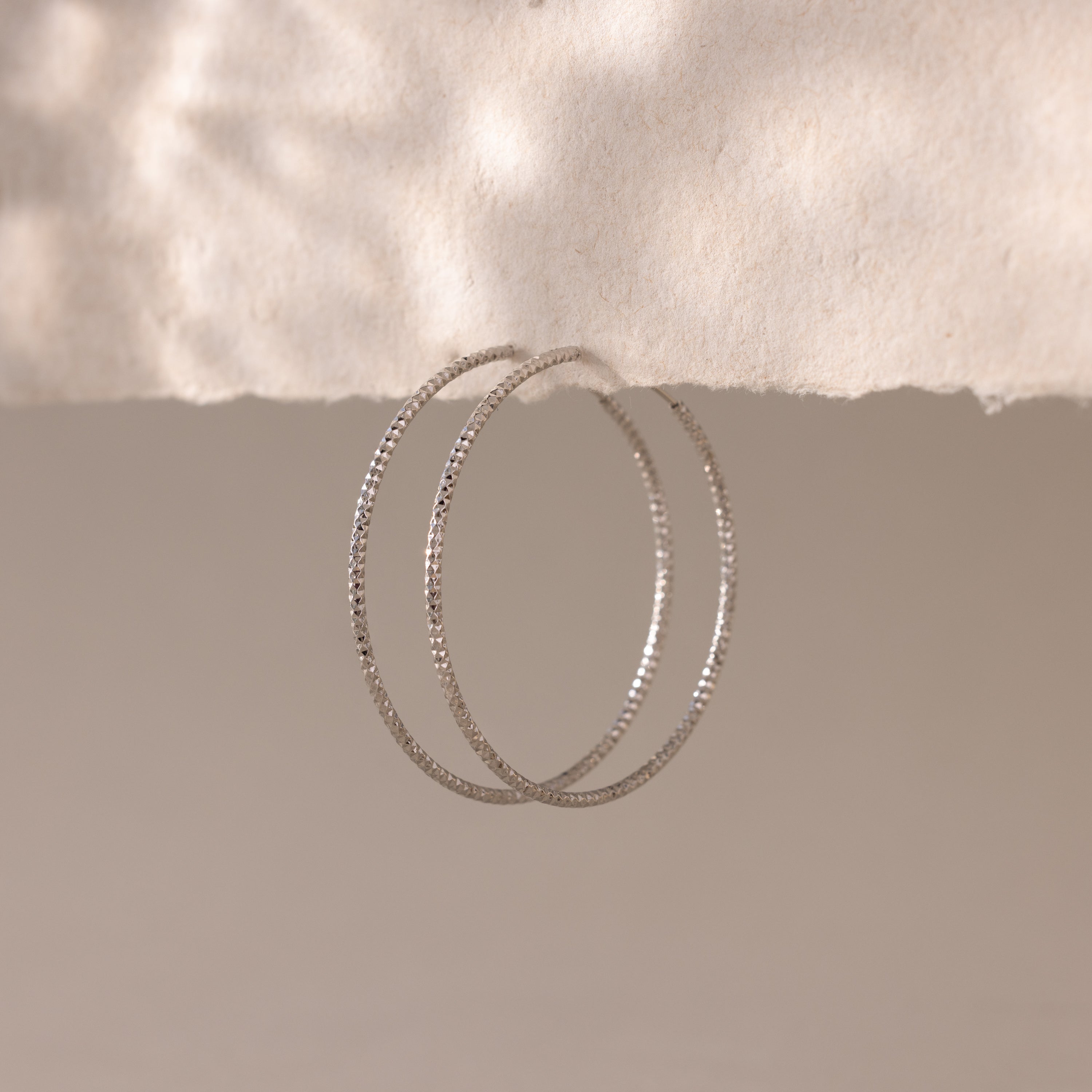 A pair of large, silver-toned hoop earrings with a faceted texture are suspended from the edge of a softly torn piece of off-white paper.