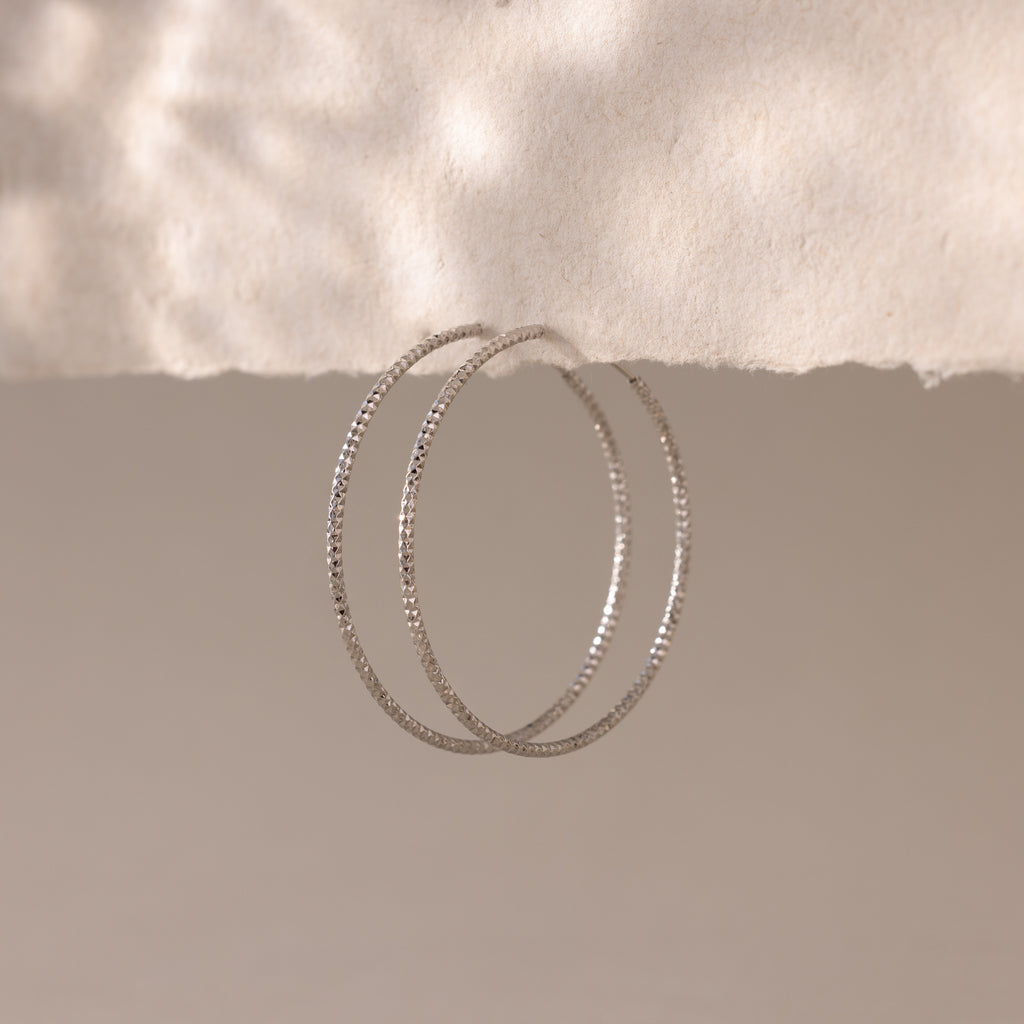 A pair of large, silver-toned hoop earrings with a faceted texture are suspended from the edge of a softly torn piece of off-white paper.