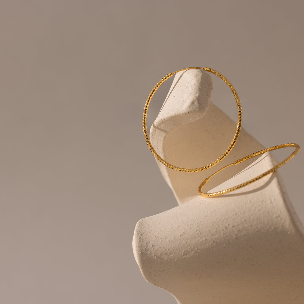A pair of textured gold hoop earrings displayed on an abstract, cream-colored sculptural stand. One hoop rests securely on the upward-curving edge of the display, while the second lies flat against the base.