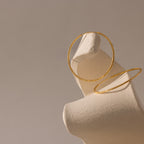 A pair of textured gold hoop earrings displayed on an abstract, cream-colored sculptural stand. One hoop rests securely on the upward-curving edge of the display, while the second lies flat against the base.