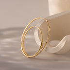 Two large, textured gold hoop earrings rest delicately on a sculptural, matte ceramic stand with soft curves. The hammered finish on the hoops catches and reflects ambient light, casting subtle shadows on the smooth surface beneath.