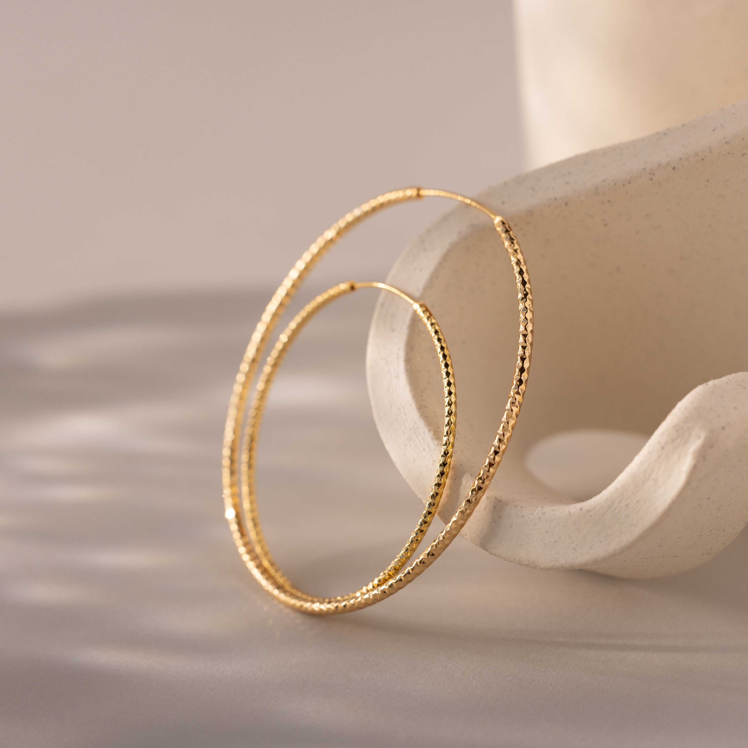 Two large, textured gold hoop earrings rest delicately on a sculptural, matte ceramic stand with soft curves. The hammered finish on the hoops catches and reflects ambient light, casting subtle shadows on the smooth surface beneath.