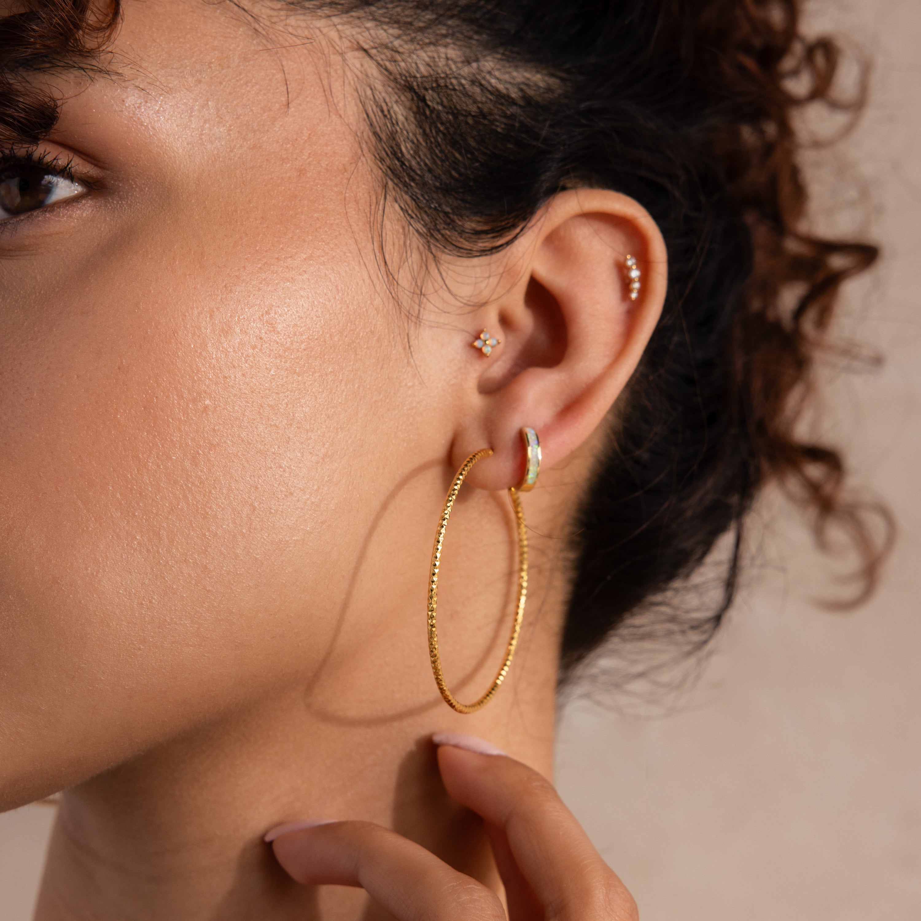 Close-up of a woman's ear adorned with multiple earrings, including a large, gold-toned faceted hoop earring as the main focal point. She also wears a small iridescent huggie, a tiny flower-shaped stud, and a delicate trio of diamond-like studs along the upper ear.