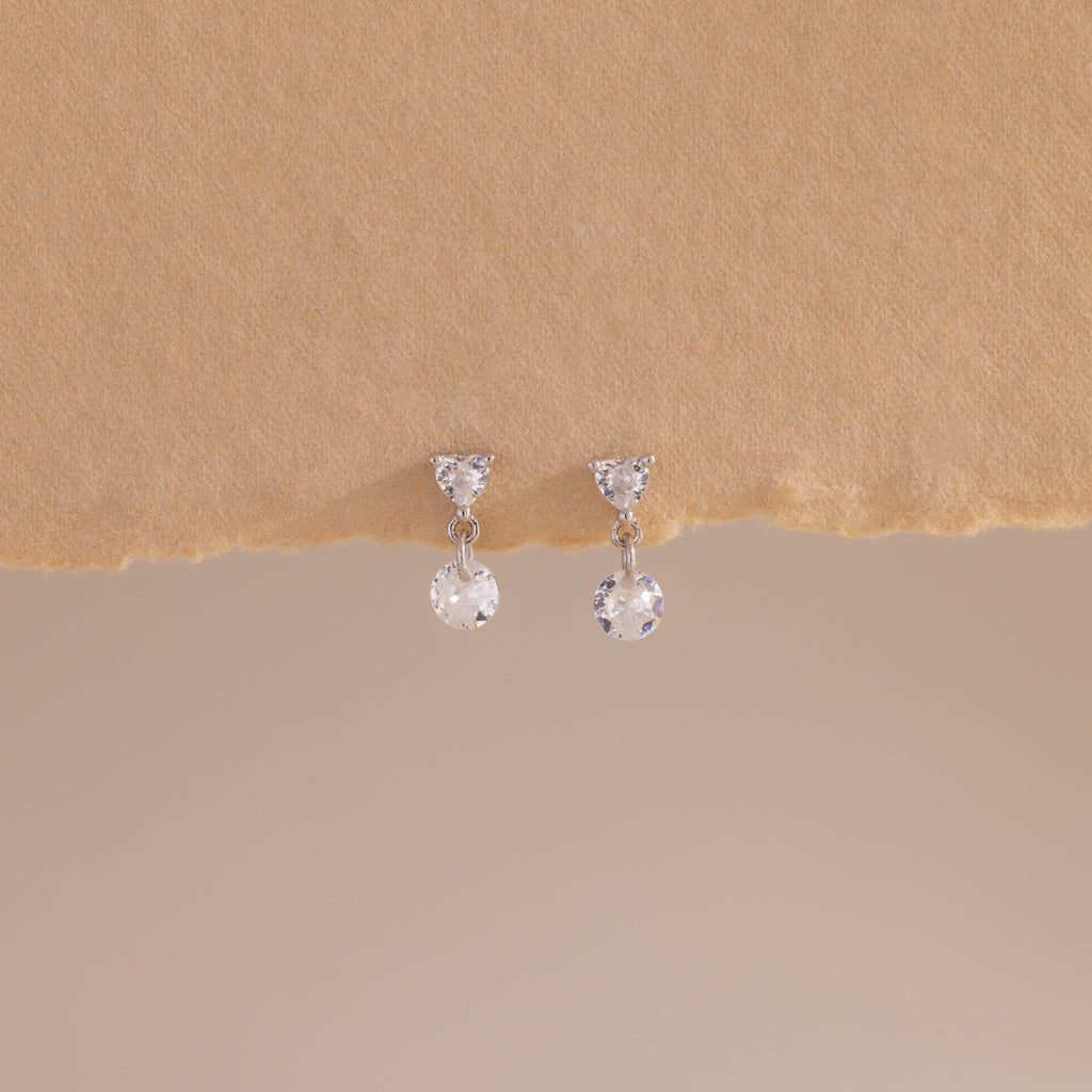 A close-up of a pair of sterling silver flat back earrings featuring heart-shaped studs with a dangling round crystal, displayed against a soft beige textured surface.