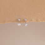A close-up of a pair of sterling silver flat back earrings featuring heart-shaped studs with a dangling round crystal, displayed against a soft beige textured surface.