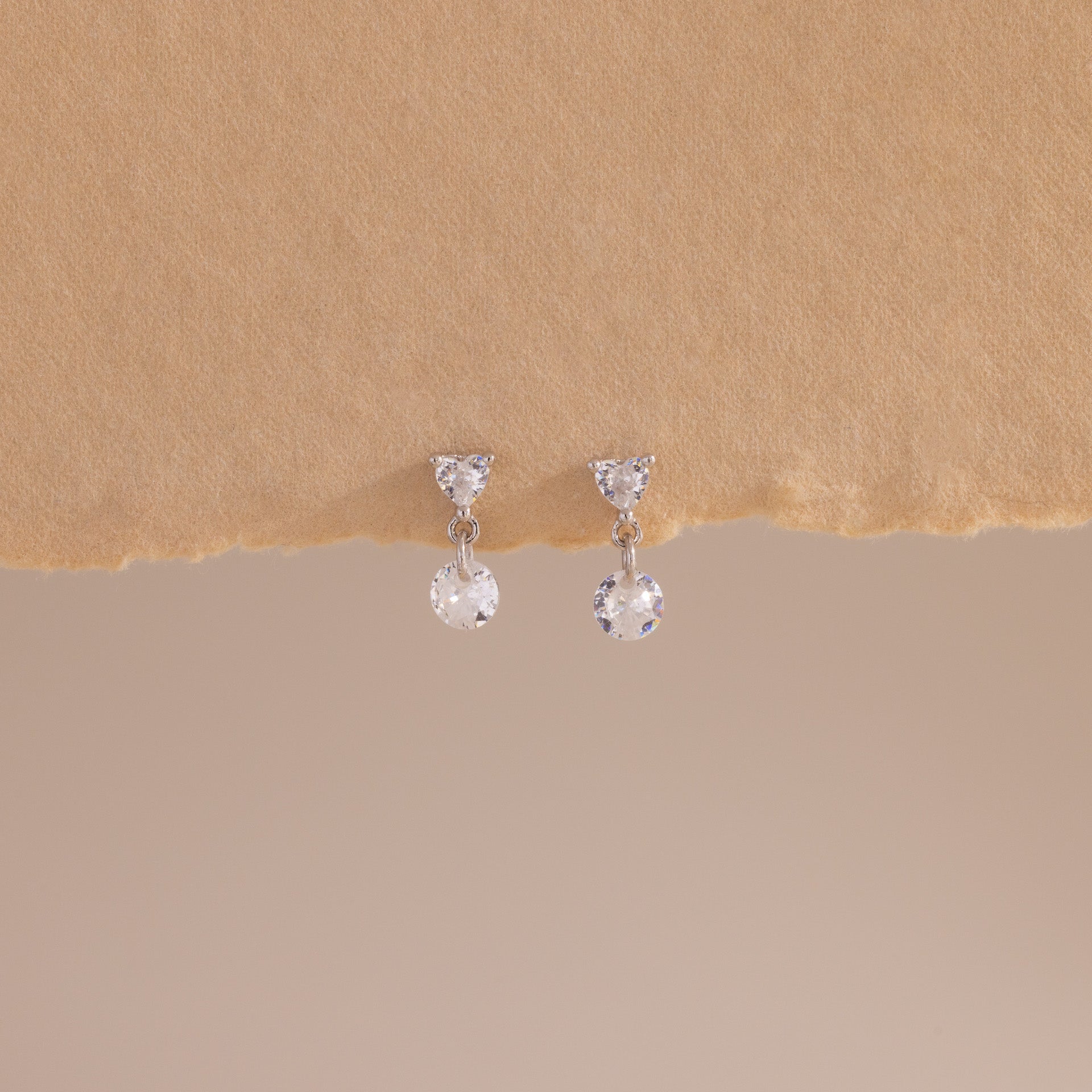A close-up of a pair of sterling silver flat back earrings featuring heart-shaped studs with a dangling round crystal, displayed against a soft beige textured surface.