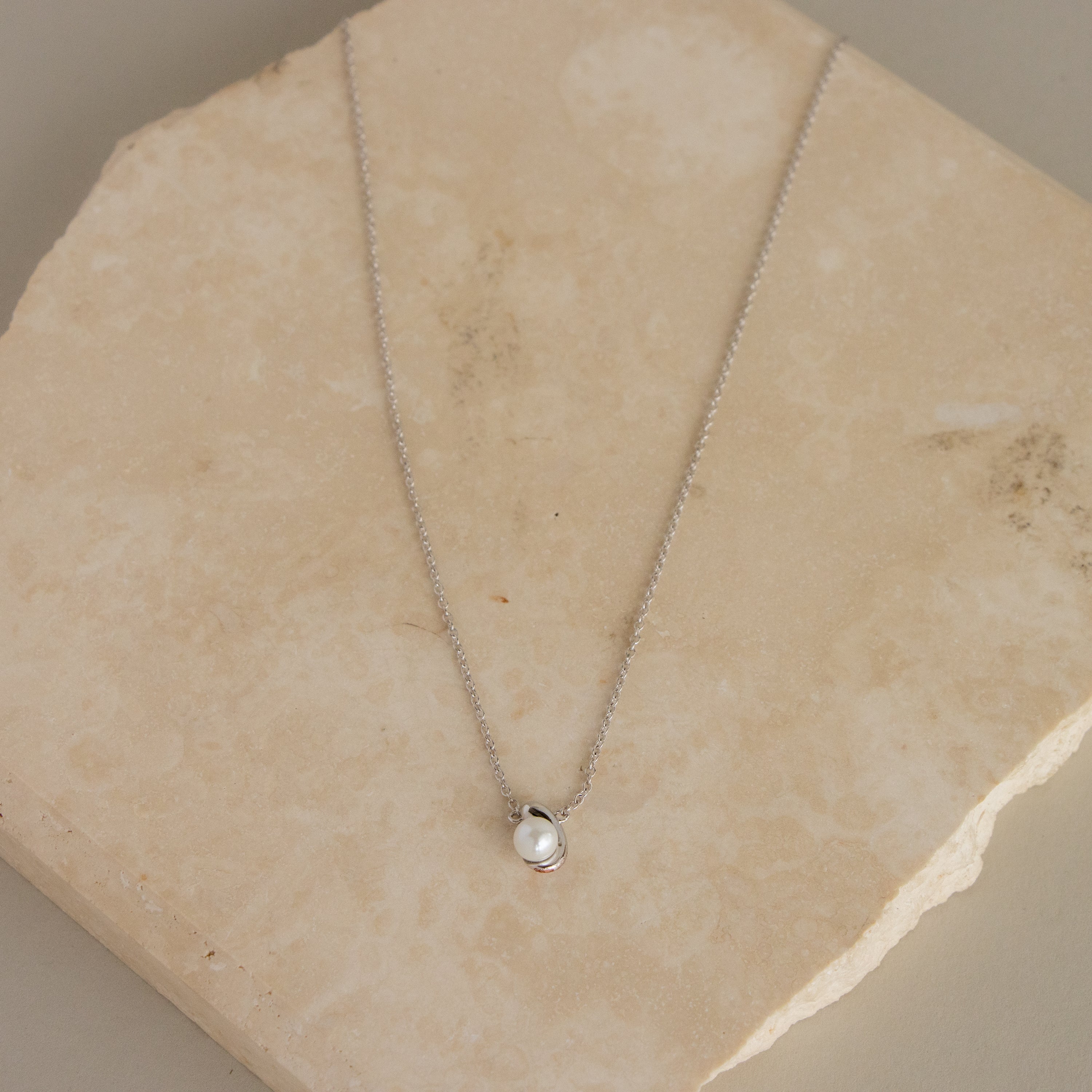 The Silver Pearl Curve Charm Necklace features a delicate silver chain with a small pearl pendant, elegantly displayed on a light stone surface.